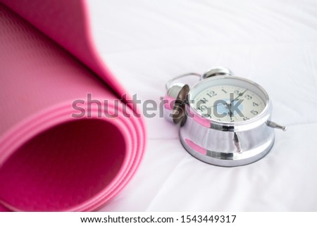 Alarm clock with healthy yoga mat on bed. A roll of yoga mat is sports gear for stretching & balance sports. The flexibility exercise is for health care & wellness. Time to workout concept.   