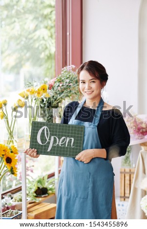 Lovely smiling Vietnamese florist in blue apron holding open sign and looking at camera
