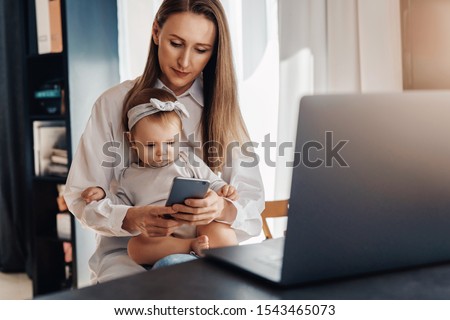 Young woman texting on smartphone with baby girl sitting on her knees and looking at screen. Mother working at home distantly using wireless connected devices for job. Freelance vacancies for parents 