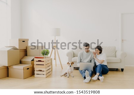 Affectionate married couple move in new home, relax on floor after unpacking things use laptop computer for searching home decoration ideas in internet their pet sits near, pose in purchased apartment Royalty-Free Stock Photo #1543519808