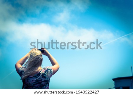 Rear view of young Caucasian lady taking photo using her smart phone. Low angle blonde girl wears shoulder strap bag use cell phone under summer cloud blue sky