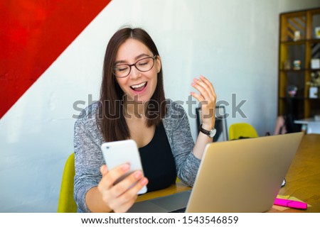 Happy smiling female in glasses skilled office worker received on mobile phone good news while sitting at workplace with open pc laptop computer 