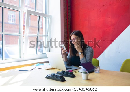 Happy smiling hipster girl in glasses received on cell telephone text message with good mood during distance work on pc laptop computer while sitting in office 