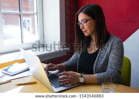 Female in casual wear and stylish glasses professional content manager searching information via pc laptop computer while sitting in office interior. Girl keyboarding on netbook, sitting in co-working