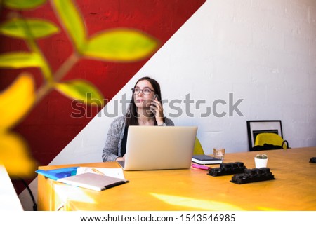 Confident female in glasses prosperous graphics designer talking via mobile phone while sitting at workplace in modern co-working space. Woman office worker having cellphone conversation  