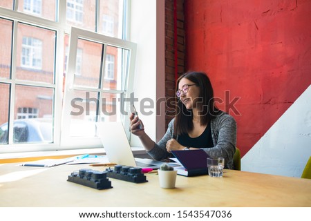 Happy smiling hipster girl in glasses reading on mobile phone text message with good news while sitting at desktop with open pc laptop computer and working tools. Female having online video call 