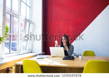 Female freelance copywriter watching video on mobile phone while sitting at workstation with pc laptop computer during distance work. Hipster girl received notifications on cell telephone 