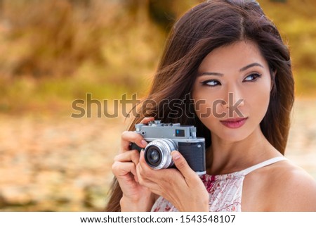 Beautiful Asian Chinese girl or young woman wearing summer dress outside in green environment taking pictures or photographs in Autumn or Fall with a retro style camera