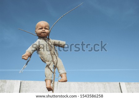 An old children's doll hanging on a branch against a concrete fence. Conceptual and metaphysical photography. Creative idea. Scary toy. Halloween