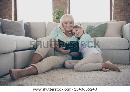 Photo of two people cute aged granny and little granddaughter sitting floor near sofa reading together girl hugging grandma listen interested story fairy tail house indoors