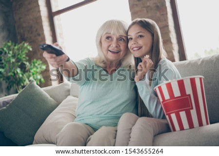 Photo of two people aged white haired granny small granddaughter sit cozy sofa watching tv eat popcorn spending summer weekend holidays together house indoors