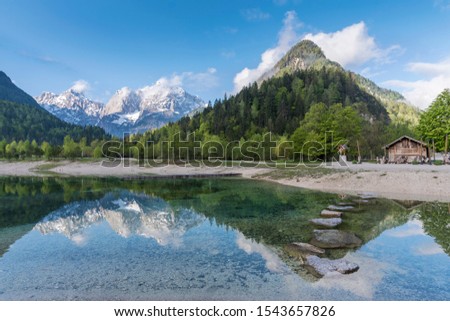Photo of lake in Kranjska Gora, Slovenia with snow covered Alps, forest and mountains in background￼￼, hut and wooden cross in foreground and reflection in lake