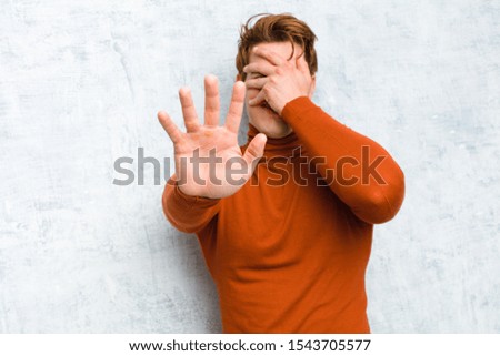 young red head man covering face with hand and putting other hand up front to stop camera, refusing photos or pictures against grunge wall