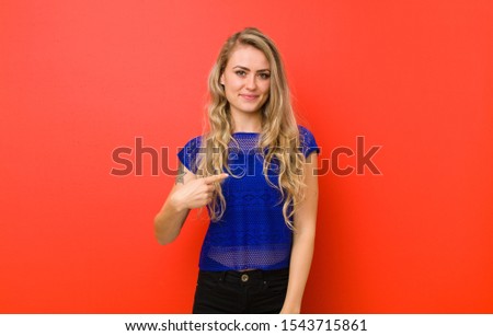 young blonde woman looking proud, confident and happy, smiling and pointing to self or making number one sign against red wall