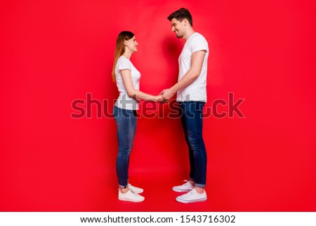 Side profile full length body size photo of cheerful positive nice couple of two people wearing jeans denim white t-shirt footwear smiling toothily holding hands isolated vivid color background