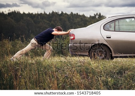 Man pushing his broken car down the country road. Driver trying to bring the car to the car service station in summer cloudy day during journey.                             