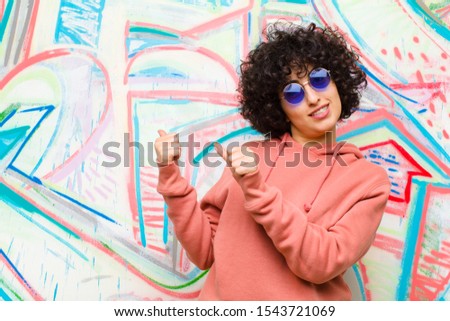 young pretty afro woman smiling cheerfully and casually pointing to copy space on the side, feeling happy and satisfied against graffiti wall