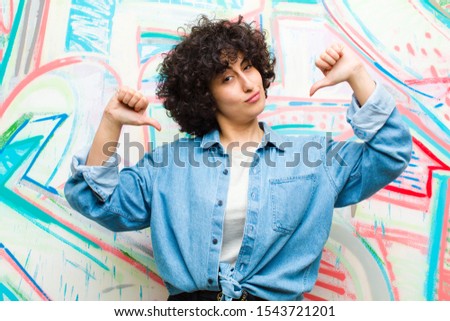 young pretty afro woman looking sad, disappointed or angry, showing thumbs down in disagreement, feeling frustrated against graffiti wall