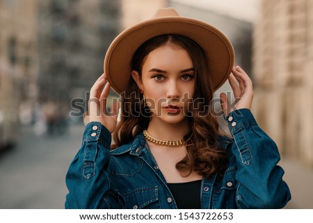 Outdoor close up fashion portrait of young elegant lady wearing beige fedora hat, trendy chain necklace, blue denim shirt, posing in street of European city. Copy, empty space for text Royalty-Free Stock Photo #1543722653