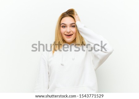 young blonde woman looking happy, astonished and surprised, smiling and realizing amazing and incredible good news against white wall