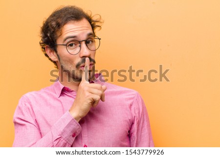 young handsome man asking for silence and quiet, gesturing with finger in front of mouth, saying shh or keeping a secret against orange wall