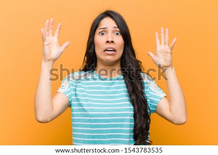 young pretty hispanic woman screaming with hands up in the air, feeling furious, frustrated, stressed and upset against brown wall