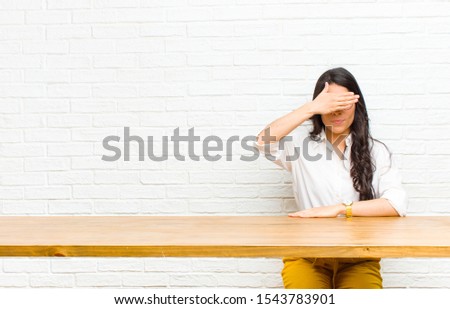 young  pretty latin woman covering eyes with one hand feeling scared or anxious, wondering or blindly waiting for a surprise sitting in front of a table