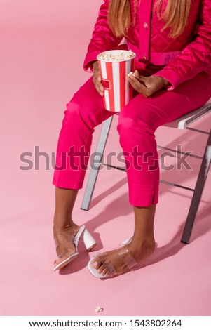 cropped view of stylsh african american woman sitting on chair with cup of popcorn on pink background, fashion doll concept