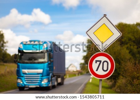 A rectangular yellow white and a round red sign on a street in Germany. It's a priority road with a speed limit of 70. A blue truck is coming.