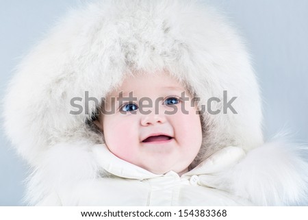 Funny sweet baby girl wearing a big fur hat and a white winter snow suit on light blue background