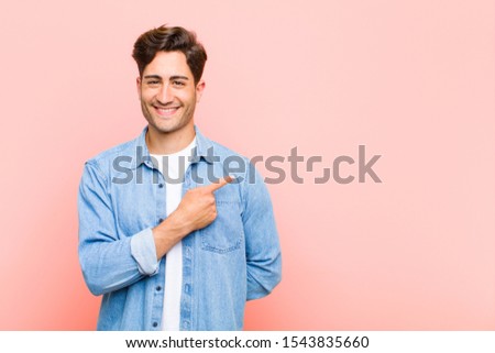 young handsome man smiling cheerfully, feeling happy and pointing to the side and upwards, showing object in copy space against pink background