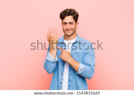 young handsome man looking impatient and angry, pointing at watch, asking for punctuality, wants to be on time against pink background