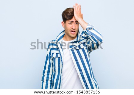 young handsome man raising palm to forehead thinking oops, after making a stupid mistake or remembering, feeling dumb against blue background