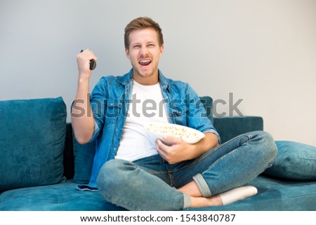 handsome young man cheers on his favorite team while watching football game eating popcorn on the sofa at home feeling happy