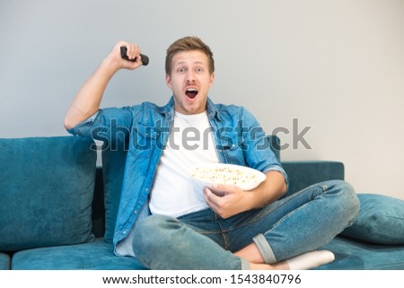 handsome young man cheers on his favorite team while watching football game eating popcorn on the sofa at home