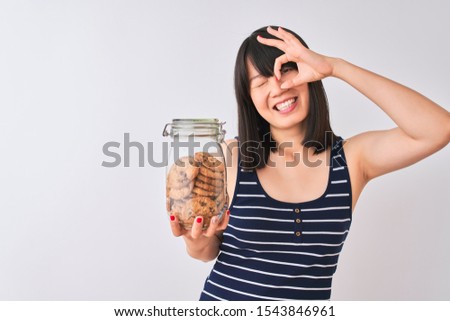 Young beautiful Chinese woman holding jar of cookies over isolated white background with happy face smiling doing ok sign with hand on eye looking through fingers