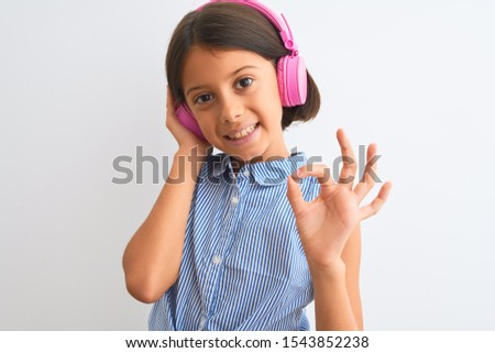 Beautiful child girl listening to music using headphones over isolated white background doing ok sign with fingers, excellent symbol