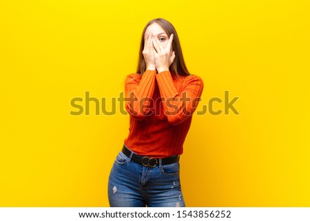 young pretty woman covering face with hands, peeking between fingers with surprised expression and looking to the side against orange background Royalty-Free Stock Photo #1543856252