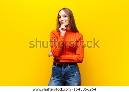 young pretty woman smiling with a happy, confident expression with hand on chin, wondering and looking to the side against orange background Royalty-Free Stock Photo #1543856264
