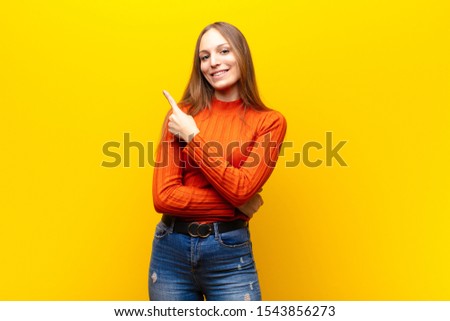 young pretty woman smiling cheerfully, feeling happy and pointing to the side and upwards, showing object in copy space against orange background Royalty-Free Stock Photo #1543856273