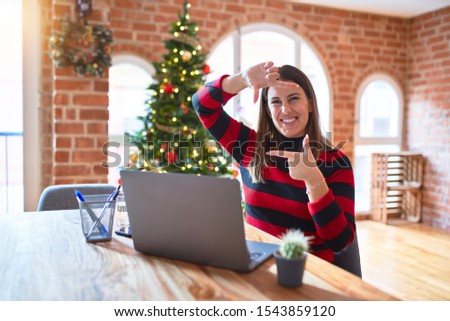 Beautiful woman sitting at the table working with laptop at home around christmas tree smiling making frame with hands and fingers with happy face. Creativity and photography concept.