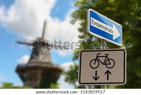 A blue and white sign shows a one way street. An additional sign allows cyclists to use both directions. In the background a windmill. Translation: one way street.
