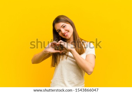 young pretty woman smiling and feeling happy, cute, romantic and in love, making heart shape with both hands against orange background Royalty-Free Stock Photo #1543866047