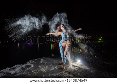 Red-haired woman in a blue dress with a deep neckline dancing in clouds of flour. A girl on the river bank jumping scattering white powder.