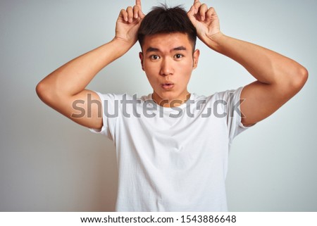 Young asian chinese man wearing t-shirt standing over isolated white background doing funny gesture with finger over head as bull horns