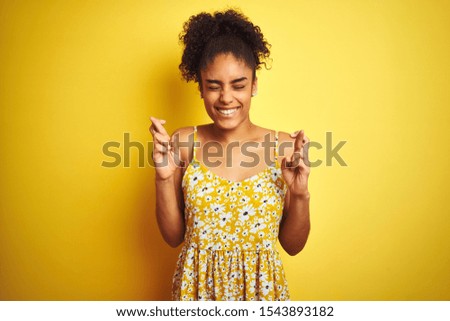 African american woman wearing casual floral dress standing over isolated yellow background gesturing finger crossed smiling with hope and eyes closed. Luck and superstitious concept.