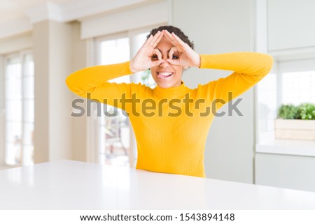 Beautiful african american woman with afro hair wearing a casual yellow sweater doing ok gesture like binoculars sticking tongue out, eyes looking through fingers. Crazy expression.