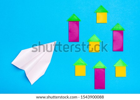 White paper plane flies over colored figures of houses on a blue cardboard background. Top view. Plane crash danger concept