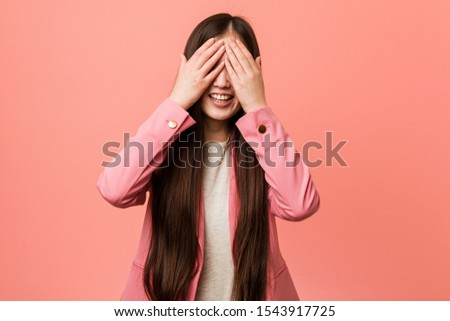 Young business chinese woman wearing pink suit covers eyes with hands, smiles broadly waiting for a surprise.