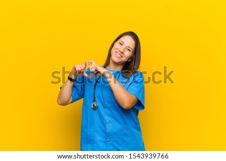 smiling and feeling happy, cute, romantic and in love, making heart shape with both hands isolated against yellow wall